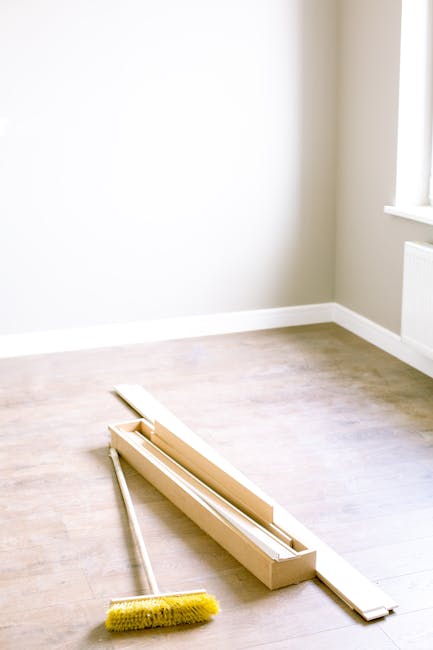 A clean, empty room with light-colored wooden flooring and beige walls illuminated by natural light from a window. In the corner, there is a wooden window frame and a white radiator below the window. On the floor, there is a yellow-handled cleaning brush and a wooden window sash removal tool resting nearby, indicating ongoing window surface cleaning or replacement. The scene reflects thorough preparation and surface cleaning, consistent with professional domestic cleaning services by Cleaners SW7, related to avoiding common end of tenancy cleaning mistakes in Kensington, SW7, focusing on detailed surface and window maintenance.