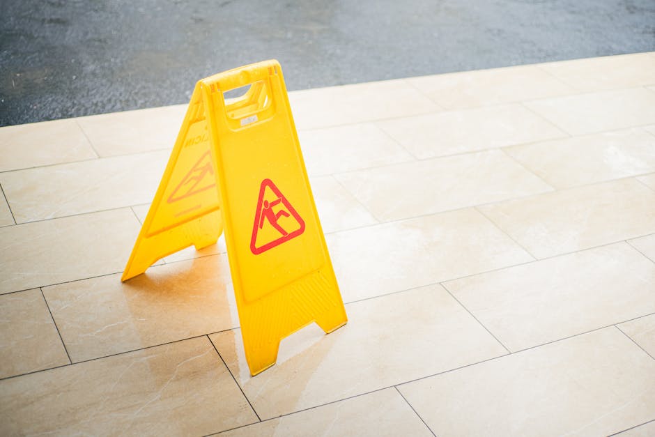 A bright yellow caution wet floor sign positioned on a beige tiled floor in an indoor or outdoor space. The sign features red warning symbols and text indicating a slip hazard. The tiles have a smooth, polished surface that appears clean and dry, with visible grout lines. The background shows a section of dark flooring or pavement, contrasting with the light-colored tiles. The scene is well-lit, emphasizing the glossy finish of the tiles, and the sign is prominently placed, suggesting routine surface cleaning or maintenance by Cleaners SW7, specialists in domestic and commercial sanitisation and deep cleaning, aimed at avoiding common end of tenancy cleaning mistakes in Kensington, SW7.