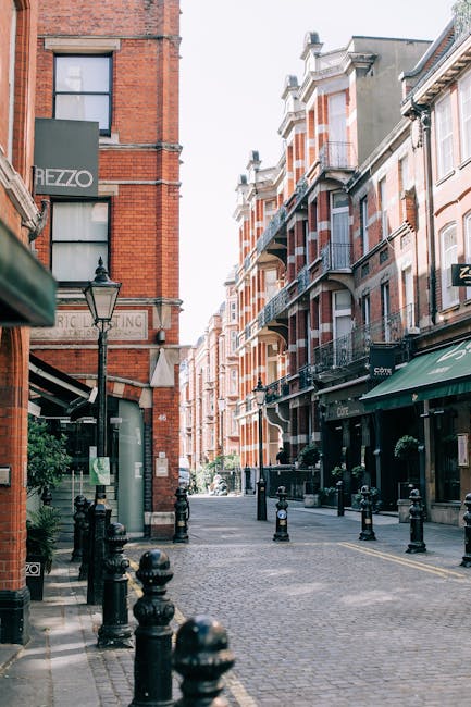Photograph of a narrow, cobblestone street in Kensington high street, SW7, lined with red-brick and white facade residential buildings featuring balconies and large windows. The scene includes a street lamp, black bollards along the pavement, and retail storefronts with green awnings. The area appears clean and well-maintained, with no visible litter or debris, and the lighting suggests it is daytime with natural sunlight illuminating the street. The image showcases an exterior urban setting, emphasizing the tidy and organized environment indicative of professional cleaning standards that Cleaners SW7 offers for both domestic and commercial spaces in the area.