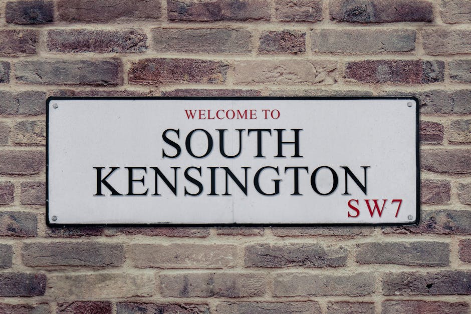 Photograph of a narrow, cobblestone street in Kensington high street, SW7, lined with red-brick and white facade residential buildings featuring balconies and large windows. The scene includes a street lamp, black bollards along the pavement, and retail storefronts with green awnings. The area appears clean and well-maintained, with no visible litter or debris, and the lighting suggests it is daytime with natural sunlight illuminating the street. The image showcases an exterior urban setting, emphasizing the tidy and organized environment indicative of professional cleaning standards that Cleaners SW7 offers for both domestic and commercial spaces in the area.