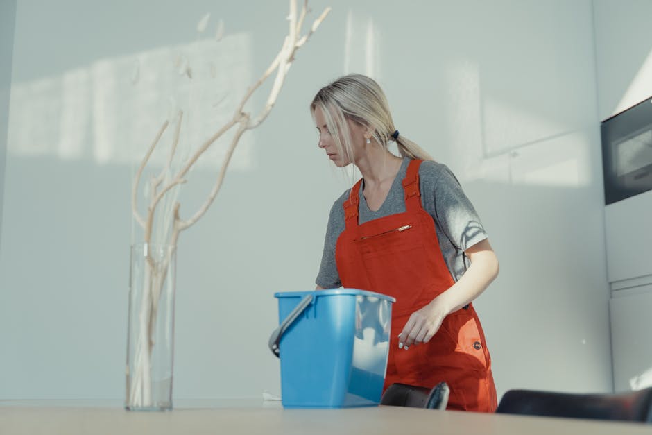 A female cleaner with blonde hair tied in a ponytail, wearing a grey T-shirt and red overalls, is engaged in surface cleaning in a modern kitchen. She is holding a blue bucket and appears to be wiping a counter or table surface. The kitchen features a white glossy wall, a ceramic tile countertop, and stainless steel appliances. Natural light illuminates the space, highlighting the tidy and hygienic condition of the surfaces. The scene reflects thorough domestic cleaning and maintenance, consistent with professional services offered by Cleaners SW7, emphasizing cleanliness and sanitisation of kitchen surfaces using appropriate tools and techniques.