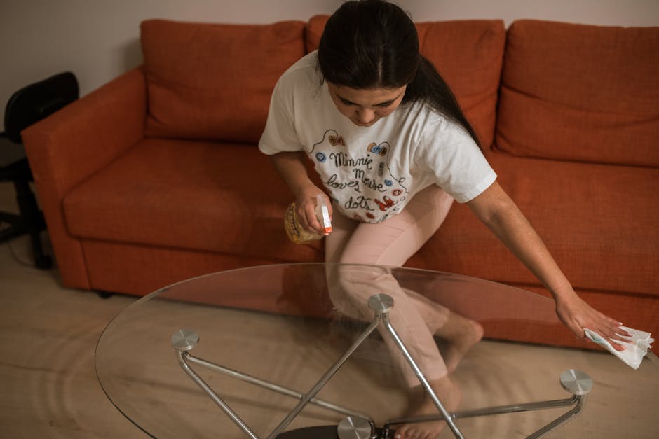 A woman is engaged in domestic cleaning, wiping a large, round glass coffee table with a cloth in a living room. She is wearing a white t-shirt adorned with colorful print designs and light pink pants. The glass surface of the table is clean and reflective, showing no dust or fingerprints. The table has a metal frame with a minimalist design, featuring a transparent, smooth surface that appears spotless and polished. Behind her, an orange fabric sofa with a textured surface is visible, placed against a plain, light-colored wall. The scene is well-lit with natural or artificial light, emphasizing the cleanliness and orderliness of the space, aligning with surface cleaning and hygiene maintenance promoted by Cleaners SW7 in their domestic cleaning services.