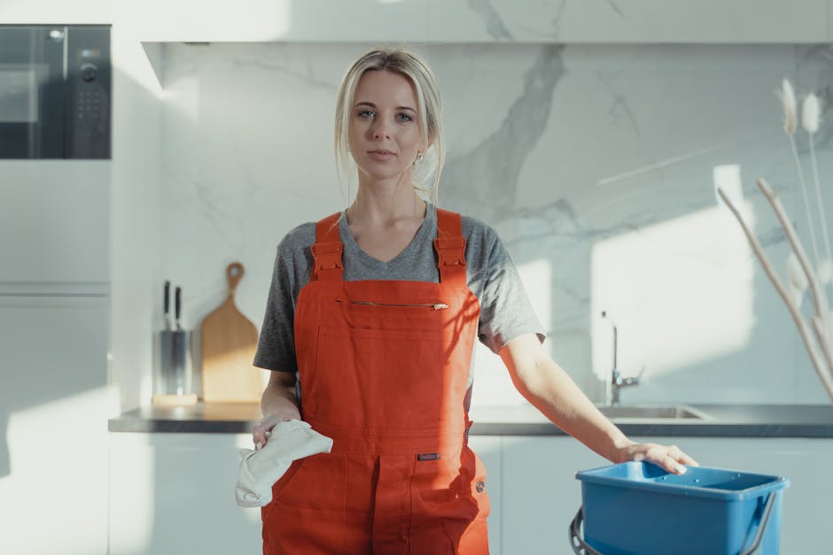 A female cleaner with blonde hair tied in a ponytail, wearing a grey T-shirt and red overalls, is engaged in surface cleaning in a modern kitchen. She is holding a blue bucket and appears to be wiping a counter or table surface. The kitchen features a white glossy wall, a ceramic tile countertop, and stainless steel appliances. Natural light illuminates the space, highlighting the tidy and hygienic condition of the surfaces. The scene reflects thorough domestic cleaning and maintenance, consistent with professional services offered by Cleaners SW7, emphasizing cleanliness and sanitisation of kitchen surfaces using appropriate tools and techniques.