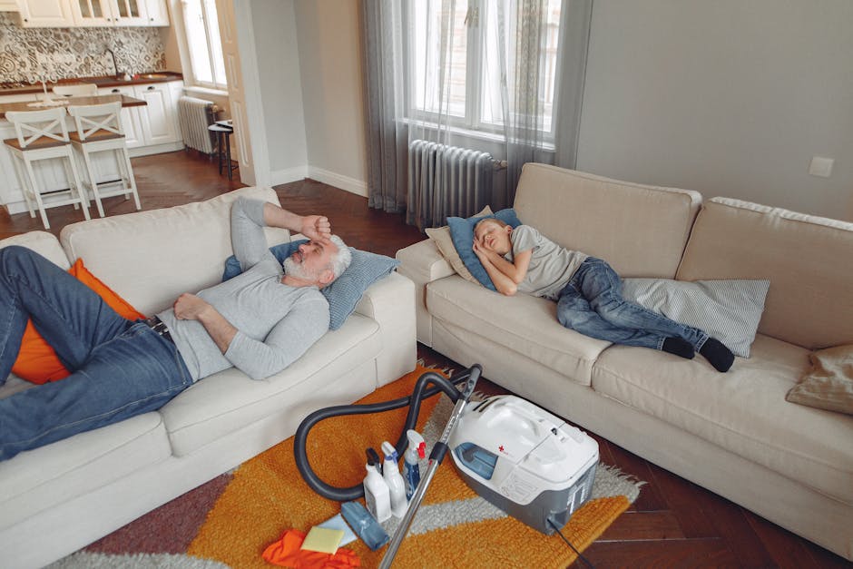 A living room scene showing two individuals resting on cream-colored sofas, with a vacuum cleaner and cleaning supplies placed on an orange and beige rug in front of them. The room features large windows with sheer curtains allowing natural light, a white radiator beneath the windows, and a wooden dining table with white chairs in the background. The surfaces appear clean and dust-free, with the sofas and floors in a well-maintained condition, illustrating a setting suitable for professional deep cleaning and sanitisation in accordance with Kensington council waste rules for carpet and sofa cleaning, as promoted by Cleaners SW7.
