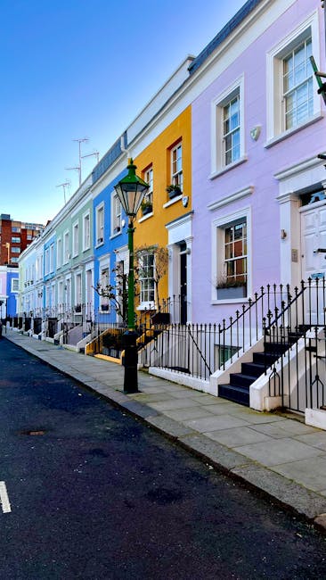 A row of colorful terraced houses on a residential street in Kensington, featuring pastel facades in shades of blue, yellow, lavender, and white. The houses have white-framed windows, black wrought-iron railings along small front steps, and decorative details around the doors. A classic black lamppost stands on the pavement, which is clean and well-maintained under bright daylight. The scene reflects a tidy, picturesque neighborhood with a vibrant, inviting atmosphere, emphasizing the importance of surface cleaning and maintenance in domestic environments, consistent with the services provided by Cleaners SW7 for end of tenancy cleaning, highlighting hygiene and presentation.
