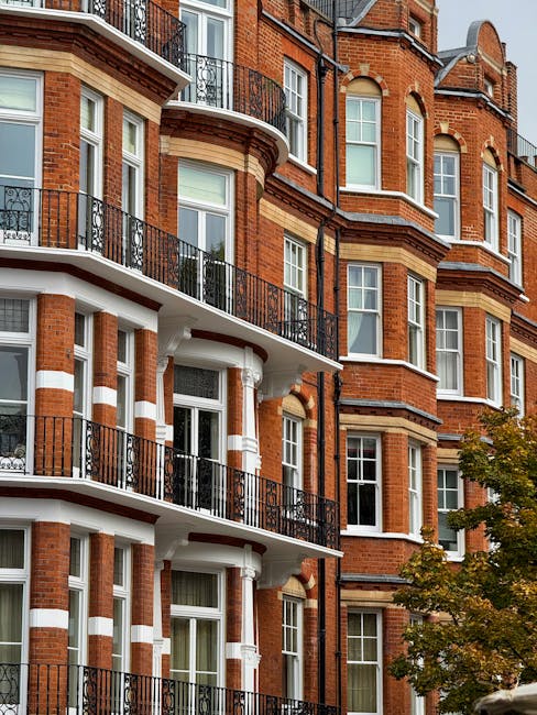 Photograph of a multi-story residential building showcasing red brick facades with white accents around the windows and balconies. The building features black wrought-iron railings on the curved balconies, which are clean and free of dirt or debris. The windows appear spotless, with clear glass reflecting natural daylight. Adjacent to the building, part of a tree with green leaves is visible, indicating an outdoor setting. The overall appearance suggests well-maintained surfaces, consistent with professional deep cleaning or surface cleaning services, as offered by Cleaners SW7, to ensure hygiene and upkeep of residential properties within the Kensington area.