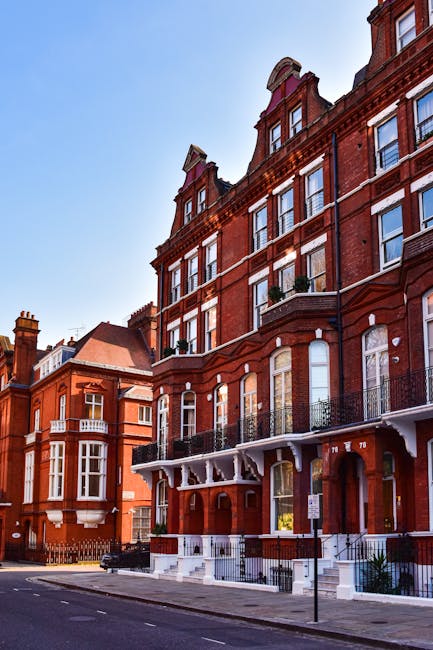 A row of colorful terraced houses on a residential street in Kensington, featuring pastel facades in shades of blue, yellow, lavender, and white. The houses have white-framed windows, black wrought-iron railings along small front steps, and decorative details around the doors. A classic black lamppost stands on the pavement, which is clean and well-maintained under bright daylight. The scene reflects a tidy, picturesque neighborhood with a vibrant, inviting atmosphere, emphasizing the importance of surface cleaning and maintenance in domestic environments, consistent with the services provided by Cleaners SW7 for end of tenancy cleaning, highlighting hygiene and presentation.