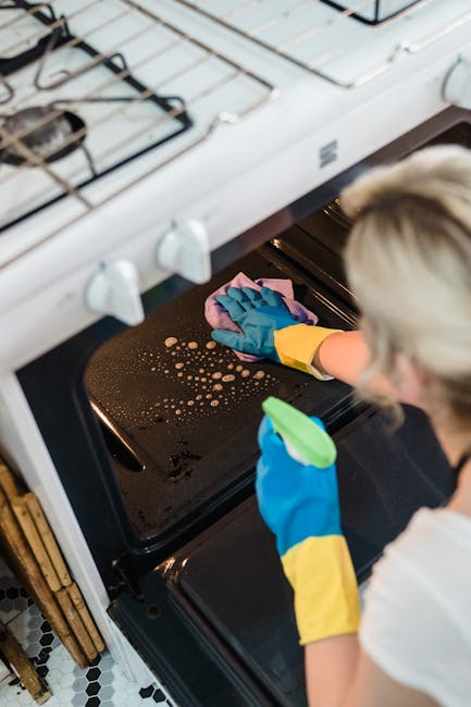 A professional cleaner from Cleaners SW7 is performing surface cleaning inside an oven in a domestic kitchen. The cleaner, wearing yellow rubber gloves and using a pink cloth, is wiping the black enamel oven door, which appears wet with cleaning solution and foam residues. The oven's metal interior and glass door reflect the surrounding light, indicating a well-lit space. Adjacent to the oven, a white gas hob with metal pan supports and control knobs is visible on a white countertop, which is clean and free of stains. The kitchen floor features black and white patterned tiles, contributing to a bright and tidy setting focused on deep cleaning and sanitisation of kitchen appliances, in line with the focused content of the Kensington oven cleaning price guide and cost breakdown, SW7, provided by [COMPANY_NAME].