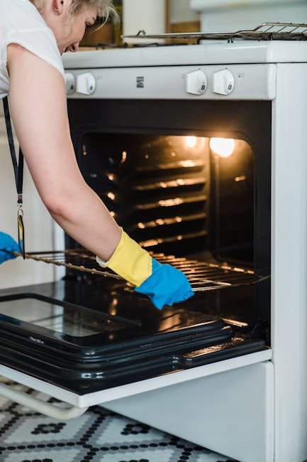 A person wearing a white shirt and yellow and blue rubber gloves is cleaning the interior of a white oven with a wire rack, inside a kitchen with a tiled black and white floor. The oven door is open, revealing the dark interior and heating elements, with a soft glow from the oven light. The countertop around the oven is clutter-free, and the scene is well-lit, highlighting the thorough cleaning process. This image showcases surface cleaning and deep cleaning practices, aligning with the services offered by Cleaners SW7 for residential kitchens, emphasizing hygiene and maintenance.
