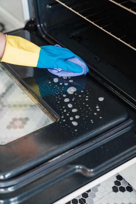 A professional cleaner from Cleaners SW7 is performing surface cleaning inside an oven in a domestic kitchen. The cleaner, wearing yellow rubber gloves and using a pink cloth, is wiping the black enamel oven door, which appears wet with cleaning solution and foam residues. The oven's metal interior and glass door reflect the surrounding light, indicating a well-lit space. Adjacent to the oven, a white gas hob with metal pan supports and control knobs is visible on a white countertop, which is clean and free of stains. The kitchen floor features black and white patterned tiles, contributing to a bright and tidy setting focused on deep cleaning and sanitisation of kitchen appliances, in line with the focused content of the Kensington oven cleaning price guide and cost breakdown, SW7, provided by [COMPANY_NAME].