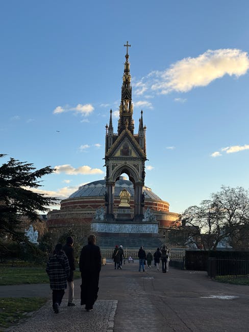 The image shows the exterior of the Royal Albert Hall with a prominent ornate spire topped by a cross, set against a bright blue sky with scattered clouds. Surrounding the monument, there are paved pathways and a few visitors walking towards the structure. Flanking the scene are trees, some with sparse foliage, indicating a possible autumn or early winter season. The area appears clean and well-maintained, with no visible litter or debris. The lighting suggests it is late afternoon, casting gentle shadows across the scene. This outdoor setting highlights the importance of maintaining the appearance of historic venues, aligning with the detailed cleaning and maintenance services offered by Cleaners SW7, as outlined in their Royal Albert Hall event cleaning service checklist.