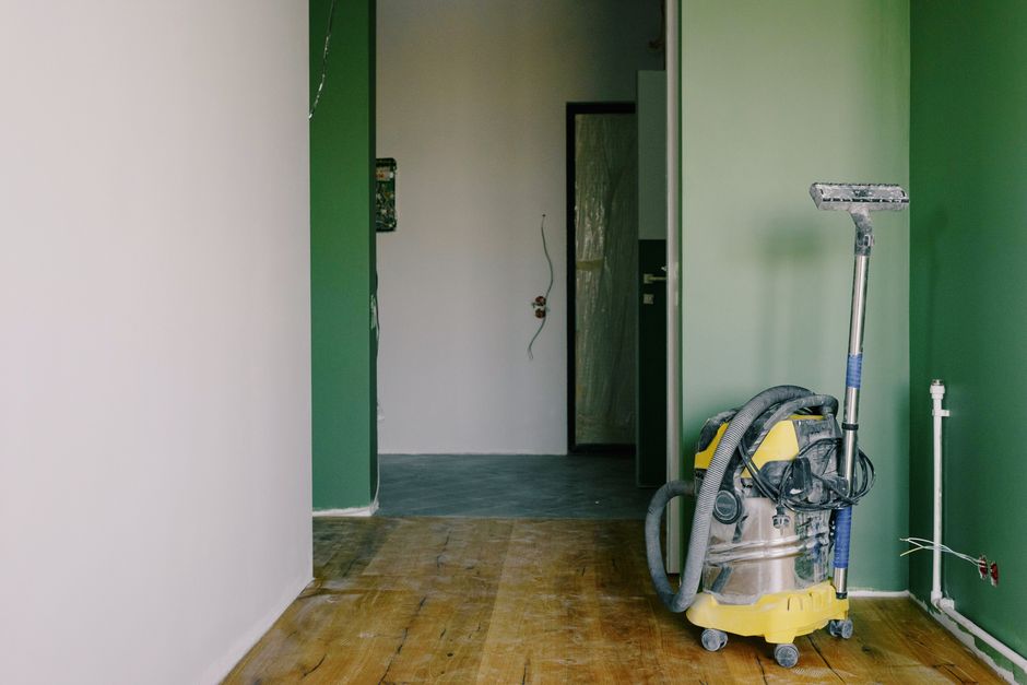The image depicts a partially renovated room with walls painted in white and green, featuring a wooden floor that appears to be in the process of cleaning. A vacuum cleaner with a yellow body and a gray hose attachment is positioned in the right foreground, indicating ongoing deep cleaning or surface sanitisation. The room contains a doorway with a black frame leading into another area, which seems to have a darker tiled floor. Visible electrical wiring on the wall suggests recent or ongoing electrical work. The lighting is natural, illuminating the surfaces and highlighting the clean, dust-free condition of the floors and walls. This image illustrates professional domestic or commercial cleaning processes, as performed by Cleaners SW7 to prepare for a property handover or end of lease cleaning.