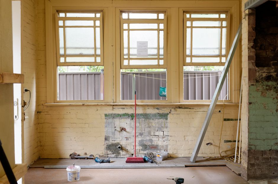 The image depicts an interior room undergoing renovation with a view of three large, rectangular windows featuring wooden grid patterns on the upper sections, allowing natural light to illuminate the space. The walls are exposed brick, with some areas showing signs of previous paint or plaster removal; the wall beneath the windows has a section of brickwork exposed after removal of plaster or drywall. A vertical metal beam leans against the right wall, and construction tools, including a red broom and a cordless drill, rest on the floor. The floor appears to be unfinished or in the process of being cleaned, with a small container of cleaning solution nearby, indicating preparation for surface cleaning or sanitisation. Outside the windows, a wooden fence and some greenery are visible, suggesting a residential setting. The space shows clear signs of ongoing renovation, with the focus on preparing surfaces for final cleaning and finishing touches, consistent with the services of Cleaners SW7 for end-of-lease or deep cleaning projects in a domestic or commercial property.